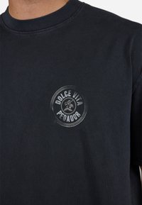 T-shirt negru din bumbac cu un imprimeu grafic rotund, estompat, care conține textul "DOLCE VITA PEGADOR" centrat pe piept. Mâneci scurte.