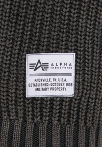 Grauer Strickstoff mit gerippter Struktur, versehen mit einem weißen Etikett, auf dem "ALPHA INDUSTRIES" und Produktdetails in schwarzer Schrift abgedruckt sind.