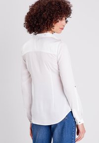 Blusa bianca a maniche lunghe con colletto, in tessuto trasparente e con polsini con bottoni, abbinata a jeans in denim blu. L'orlo ha un finish arrotondato.