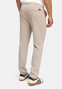 Man wearing beige slim-fit pants and white sneakers with black stripes, standing against a plain white background.