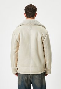 Krämfärgad shearling-jacka med en bred krage, synliga sömdetaljer och sidoglidser. Bärs över mörka jeans.