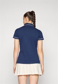 Marineblauw poloshirt met witte kraag en mouwaccenten, aangesloten ontwerp, gecombineerd met een geplooide beige rok, gezien vanuit de achterkant.