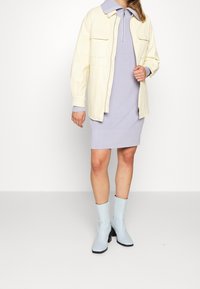 Giacca color crema con due tasche frontali sopra un vestito a coste lavanda. Stivaletti alla caviglia lucidi di colore azzurro, con punta quadrata e tacco a blocco.