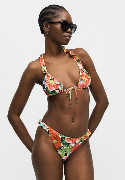 Γυναίκα με φλοράλ μπικίνι με βολάν στα τελειώματα και μαύρα γυαλιά ηλίου, ποζάρει με τα χέρια στη λεκάνη σε ουδέτερο φόντο.