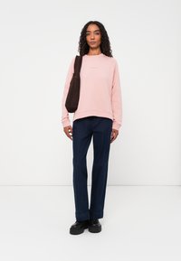 Felpa rosa chiaro con polsini a coste, abbinata a pantaloni larghi blu scuro con cucitura dritta. Gli accessori includono una borsa a tracolla marrone.