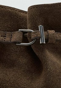 Material de ante marrón con una textura suave. Presenta una hebilla de metal plateado y una correa para el cierre. Enfocarse en los detalles y la costura.