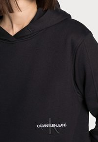 Sudadera con capucha negra hecha de tela suave. Presenta un logo bordado en blanco y una capucha texturizada. Diseño simple con un ajuste relajado.