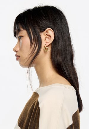 Pendientes de aro de oro con un pequeño acento circular, colgando de un cierre de metal texturizado; modelo con cabello lacio y oscuro, vista de perfil.