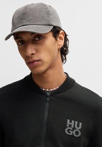Gorro gris con "HUGO" bordado en la parte frontal, que se lleva con una chaqueta negra con cremallera que muestra un logo. Cuentas ligeras alrededor del cuello.