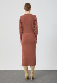 Robe longue en tricot couleur rouille avec des manches longues, taille ajustée et jupe droite ; portée avec des bottes beige jusqu'aux genoux.