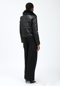 Veste en cuir noir avec un design matelassé, dotée d'un col en fausse fourrure, de poignets côtelés et d'une coupe courte. Portée avec un large pantalon noir.