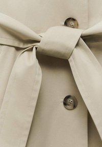 Material bej pentru trench coat, cu detaliu de curea legată. Prezintă nasturi mari și o textură netedă. Conceput cu o siluetă clasică.