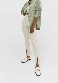 Light green suede shirt jacket, beige cropped top and fitted pants with side slits, paired with white loafers featuring gold hardware.
