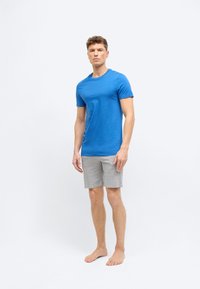 Άνδρας όρθιος ξυπόλητος, φορώντας μπλε μπλουζάκι και γκρι σορτς, με φόντο απλό λευκό, κοιτάζει μπροστά με ουδέτερη έκφραση.