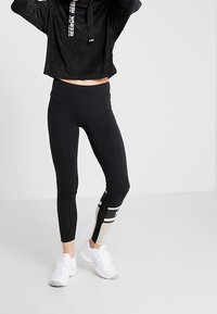 Hoodie noir court avec cordons, associé à un legging noir taille haute avec un panneau latéral beige et noir. Chaussures de sport blanches.