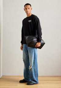 Mann in schwarzem Sweatshirt und ausgefransten weit geschnittenen Jeans, der eine große schwarze Ledertasche hält, steht auf einem Holzboden vor einer schlichten Wand.
