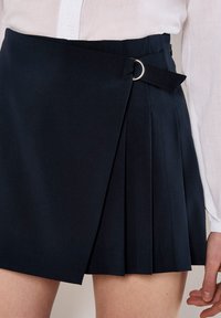 Gonna plissettata blu navy con design a incrocio, dotata di una fibbia a forma di anello argentato e una cintura in tessuto. Realizzata in materiale liscio e strutturato.