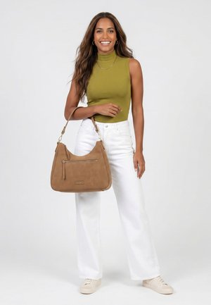Lächelnde Frau mit langen Haaren trägt ärmelloses grünes Top, weiße Hose, beige Turnschuhe und hält eine beige Handtasche mit vorderer Reißverschlusstasche.