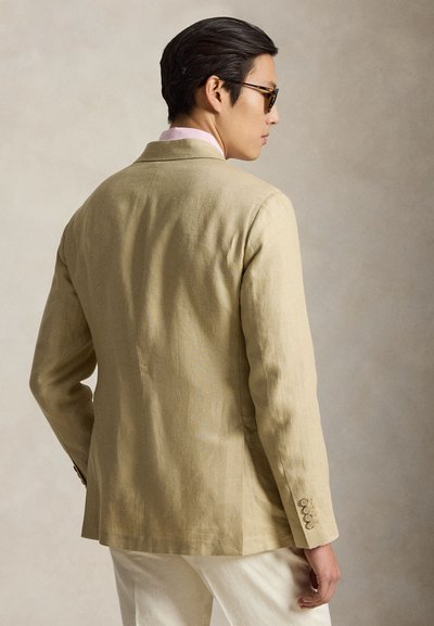 Homme portant un blazer beige, une chemise rose clair, un pantalon blanc et des lunettes de soleil, debout de profil devant un fond clair uni.