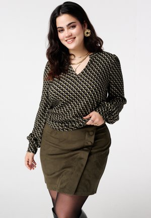 Glimlachende vrouw met donker haar draagt een top met geometrisch patroon en lange mouwen, een olijfgroene omslagrok met knopen, gouden sieraden en zwarte panty's.