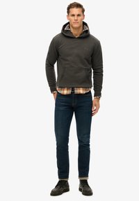 Sudadera gris con capucha y bolsillo canguro, superpuesta sobre una camisa de franela a cuadros, combinada con jeans oscuros y zapatos negros.