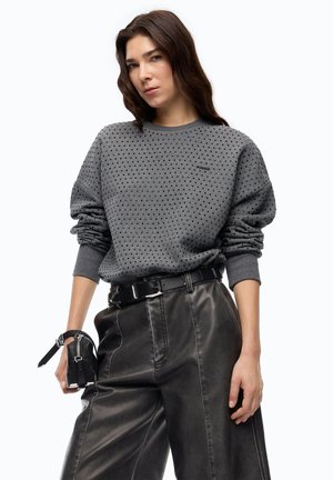 Donna che indossa una felpa grigia a pois e pantaloni neri in pelle a vita alta, che tiene una piccola pochette nera con zip nella mano sinistra.