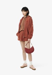 Ensemble en hoodie rouge rouille et shorts, top marron à motifs, associé à un sac à main bordeaux texturé et des baskets rose pâle avec des chaussettes blanches.