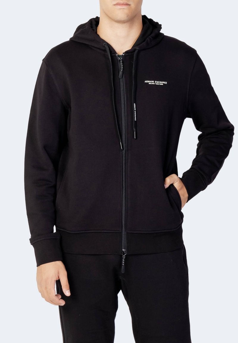 Homme en sweat à capuche noir zippé avec poches avant et logo "Armani Exchange Milano / New York" sur le côté gauche de la poitrine.