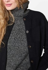 Zwarte gebreide cardigan met vijf bruine knopen over een donkergrijze turtleneck trui, met een textuur gebreid patroon.