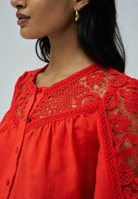 Blusa roja con intrincados detalles de encaje en los hombros, presenta un cierre de botón y tela recogida para un ajuste relajado.