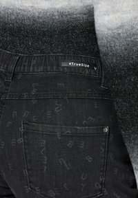 Černé džínové kalhoty s jemnými vzory písmen, pružným pasem a štítkem „#TrueSize“ na zadní straně. Mají klasické zadní kapsy.