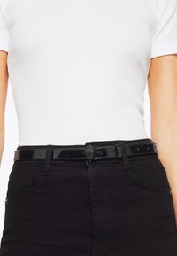 Ce ceinture en cuir verni noir avec une finition brillante présente une boucle décorative avec un design de main. Conçue pour passer à travers des passants de ceinture standards.