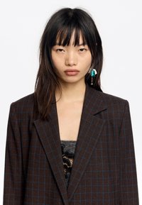 Blazer marrón a cuadros con líneas azules, que presenta solapas marcadas y hombros estructurados. Llevado sobre una blusa de encaje gris, mostrando un detalle texturizado.