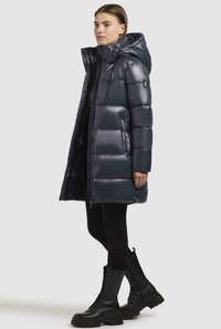 Giacca puffer blu navy con finitura lucida, cappuccio e bottoni a pressione. Abbinata a leggings neri e stivali robusti con lacci.
