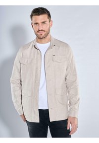 Hellbeige Hemd mit Knopfleiste, Kragen, zwei vorderen Taschen, glatter Textur und langen Ärmeln, getragen über einem weißen T-Shirt.