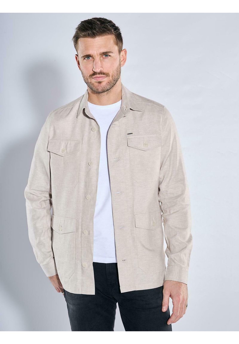 Hellbeige Hemd mit Knopfleiste, Kragen, zwei vorderen Taschen, glatter Textur und langen Ärmeln, getragen über einem weißen T-Shirt.