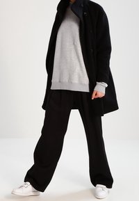 Abrigo negro, sudadera gris con bolsillo, pantalones negros holgados y zapatillas blancas. La tela parece suave; el abrigo tiene botones.
