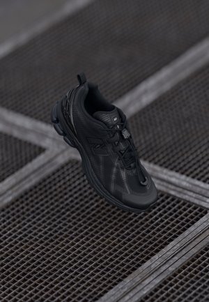 Black athletic sneaker with laces and a pull tab, positioned above metal grating in an urban setting.