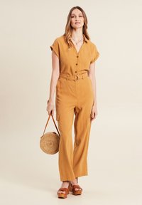 Mosterdkleurige jumpsuit met korte mouwen, knoopsluiting aan de voorkant en een ceintuur om de taille. Heeft wijde pijpen en een zak. Model houdt een ronde strooien tas vast.
