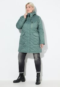 Cappotto imbottito verde, di lunghezza media, con colletto alto, chiusura con zip e due ampie tasche frontali. Indossato con jeans scuri e stivaletti neri.