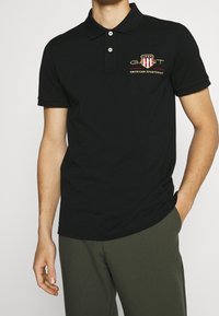 Svart poloshirt med krage med tre knappar och broderad GANT-logotyp i röd, vit och guld. Tillverkad av bomull med korta ärmar.