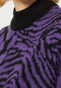 Pull rayé violet et noir avec un col haut texturé noir et un matériau tricoté duveteux présentant un motif ressemblant à celui d'un tigre.