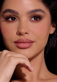 Zbliżenie twarzy o gładkiej, opalonej skórze, pełnych matowych warg w odcieniu mauve, ciemnych, wyrazistych brwiach i długich rzęsach, otoczonej czarnymi włosami.