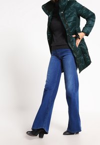 Pohodlný zelený kabát s vysokým límcem, doplněný o tmavě modré rozšířené džíny a černé boty na podpatku. Kabát má patentky a lesklou texturu.