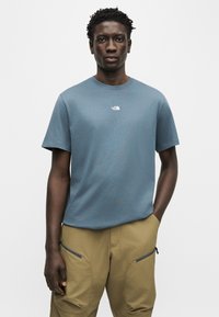 Άνδρας που φοράει μπλε κοντομάνικη μπλούζα με μικρό λευκό λογότυπο και μπεζ παντελόνι cargo με τσέπες με φερμουάρ, στέκεται με το ένα χέρι στην τσέπη.