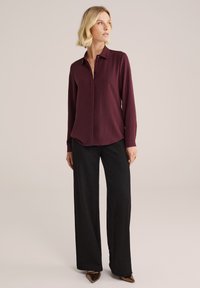 Camicia a maniche lunghe color marrone con colletto, chiusura frontale con bottoni e orlo leggermente curvo, abbinata a pantaloni neri a gamba larga e scarpe con punta.