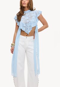 Femme portant un crop top en dentelle bleu clair avec des manches volantées et un pantalon blanc taille haute à jambes larges avec des détails de boutons.