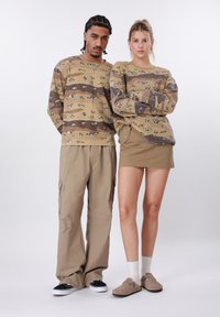 Δύο μοντέλα φορούν ταιριαστά πουλόβερ καμουφλάζ σε μπεζ και καφέ χρώματα· το ένα με παντελόνι cargo και μαύρα αθλητικά, το άλλο με μίνι φούστα και μπεζ παπούτσια χωρίς κορδόνια.