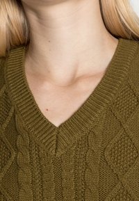 Close-up of a person’s neck and upper chest wearing an olive-green cable knit V-neck sweater.