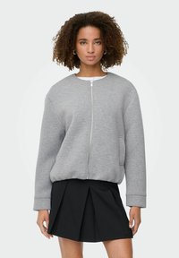 Sweatshirt gris zippé en tissu doux avec un col rond, des épaules tombantes et des poches latérales, associé à une jupe plissée noire.
